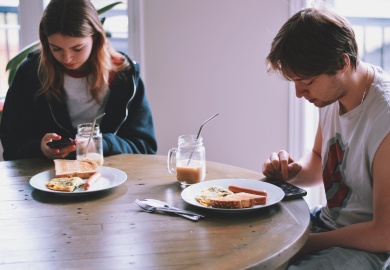 Young man and girl, sitting at kitchen table with plates in front of them and using mobile phones