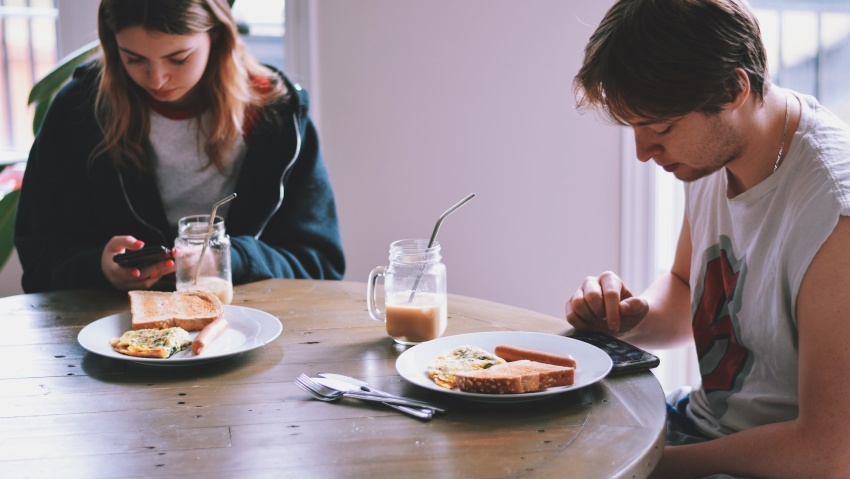 Young man and girl, sitting at kitchen table with plates in front of them and using mobile phones