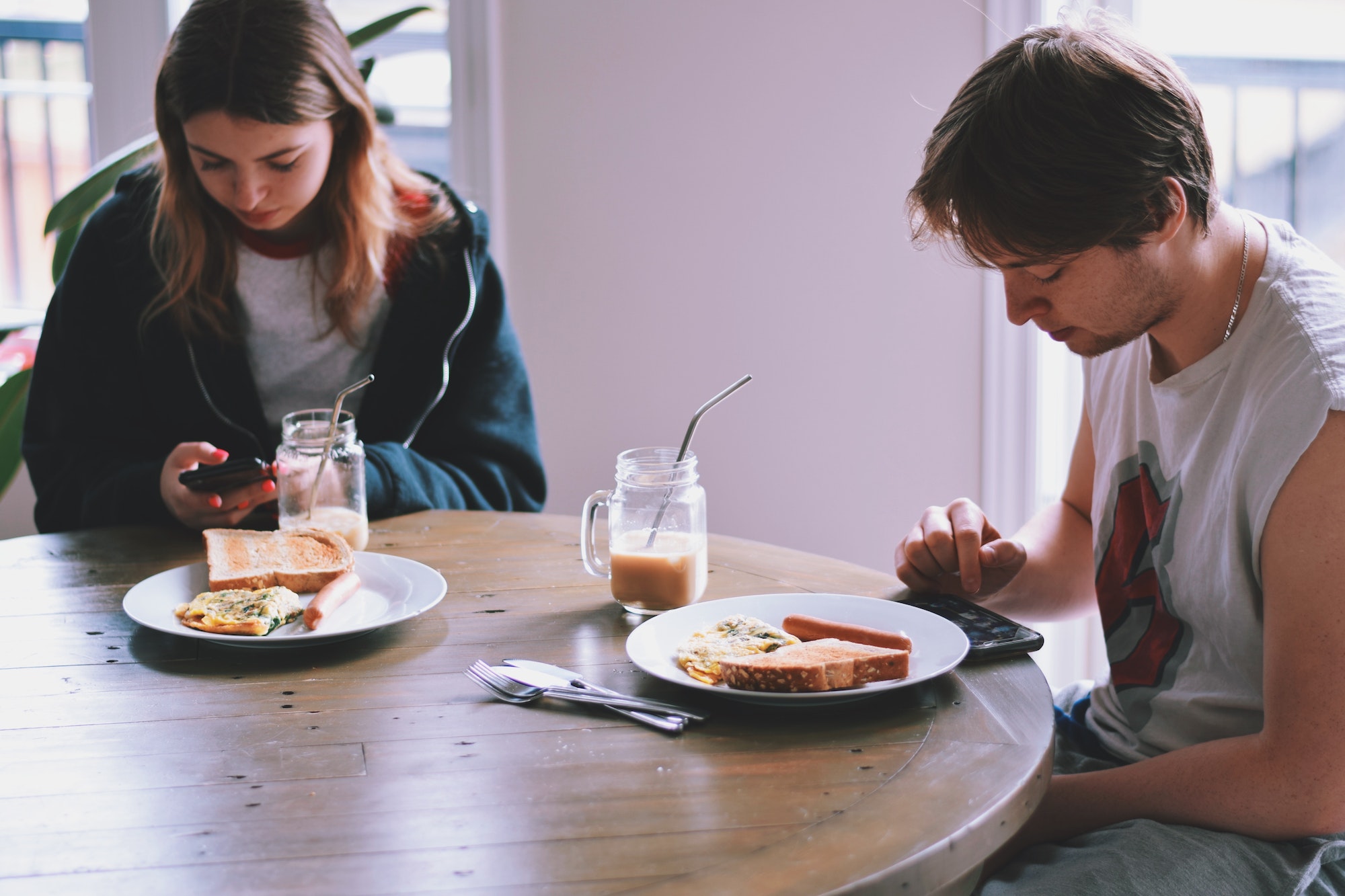 Young man and girl, sitting at kitchen table with plates in front of them and using mobile phones